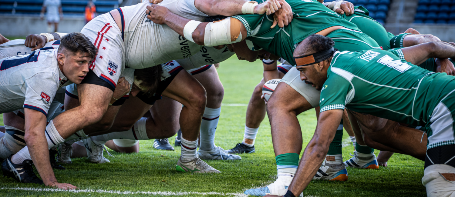 Two rugby teams engage in a scrum during a match, with players in white and green jerseys pushing against each other on the field.