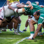 Two rugby teams engage in a scrum during a match, with players in white and green jerseys pushing against each other on the field.