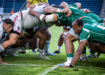 Two rugby teams engage in a scrum during a match, with players in white and green jerseys pushing against each other on the field.