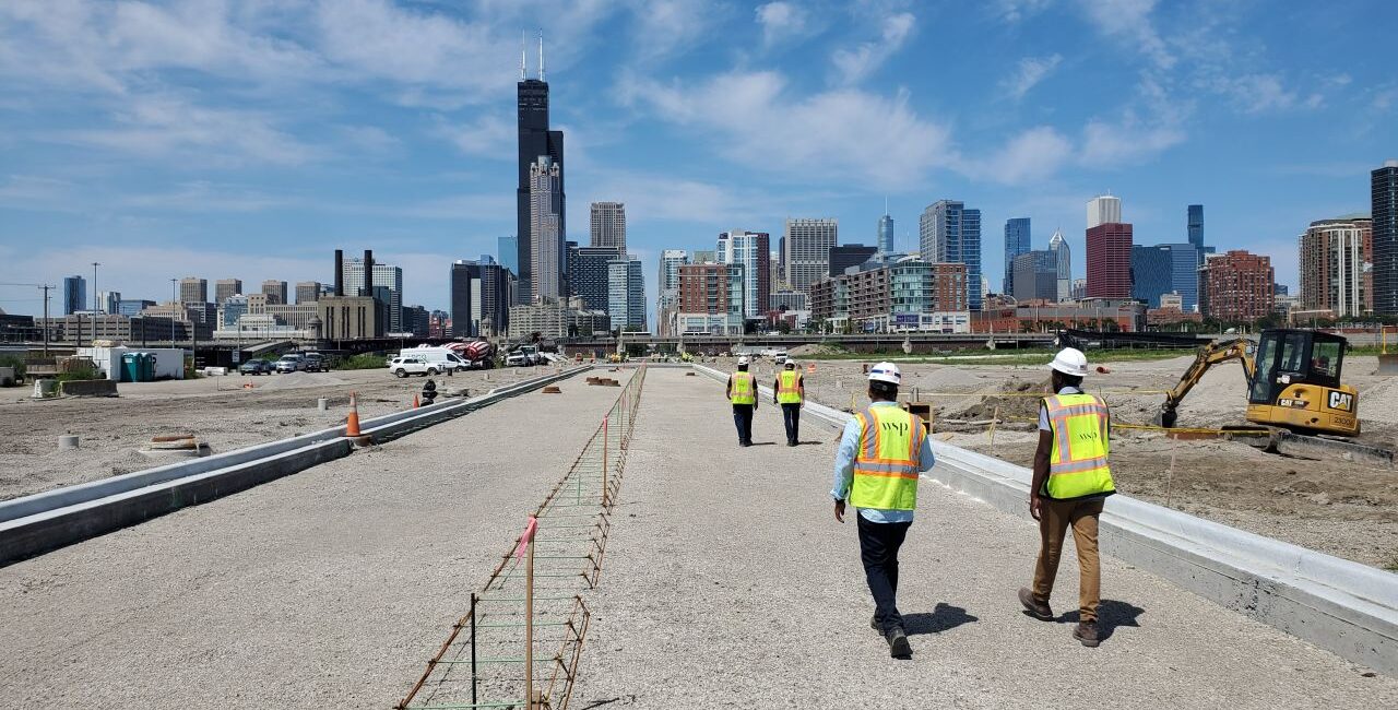Construction workers walk along a newly built roadway toward the Chicago skyline under a bright blue sky.