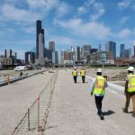Construction workers walk along a newly built roadway toward the Chicago skyline under a bright blue sky.