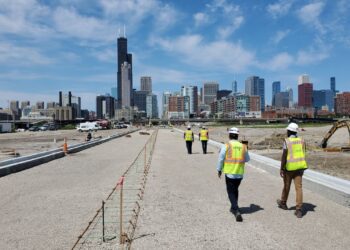 Construction workers walk along a newly built roadway toward the Chicago skyline under a bright blue sky.