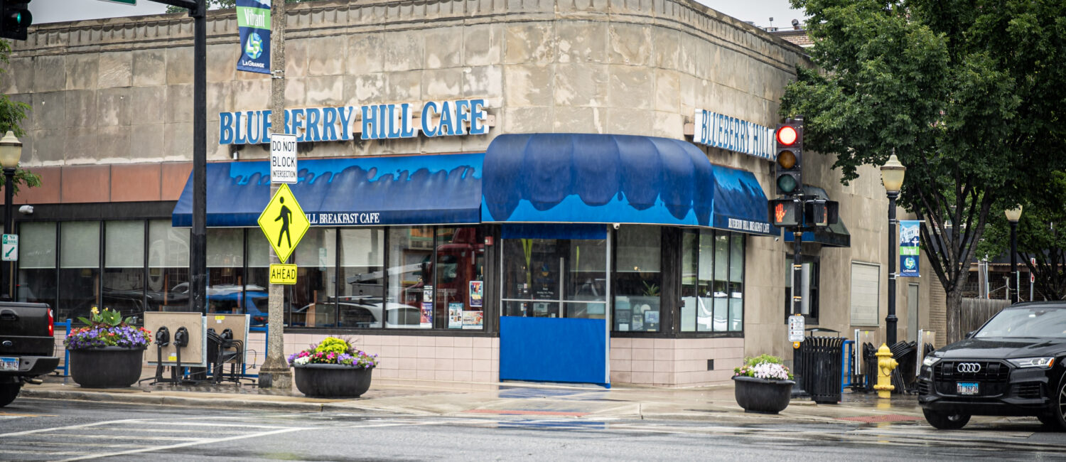 A corner view of Blueberry Hill Cafe on a rainy day, with blue awnings, wet streets, a red traffic light, crosswalk signs, and flower planters at the intersection.