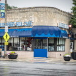 A corner view of Blueberry Hill Cafe on a rainy day, with blue awnings, wet streets, a red traffic light, crosswalk signs, and flower planters at the intersection.