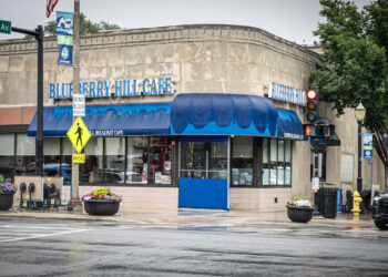 A corner view of Blueberry Hill Cafe on a rainy day, with blue awnings, wet streets, a red traffic light, crosswalk signs, and flower planters at the intersection.