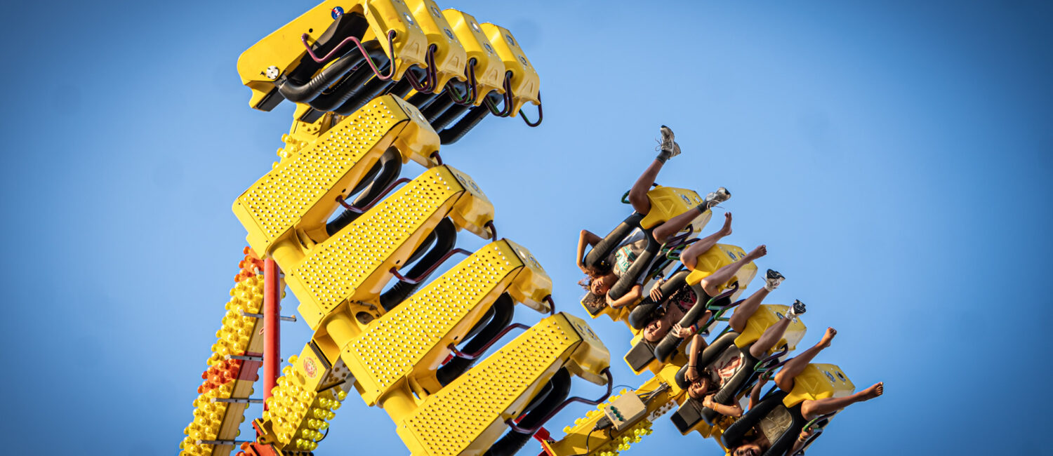 A group of people ride a yellow amusement park ride that swings high into the air against a clear blue sky. Riders are secured in seats along the ride’s arms, with several raising their legs as the ride tilts upward.