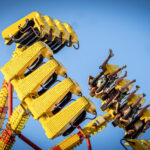 A group of people ride a yellow amusement park ride that swings high into the air against a clear blue sky. Riders are secured in seats along the ride’s arms, with several raising their legs as the ride tilts upward.