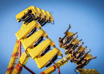 A group of people ride a yellow amusement park ride that swings high into the air against a clear blue sky. Riders are secured in seats along the ride’s arms, with several raising their legs as the ride tilts upward.
