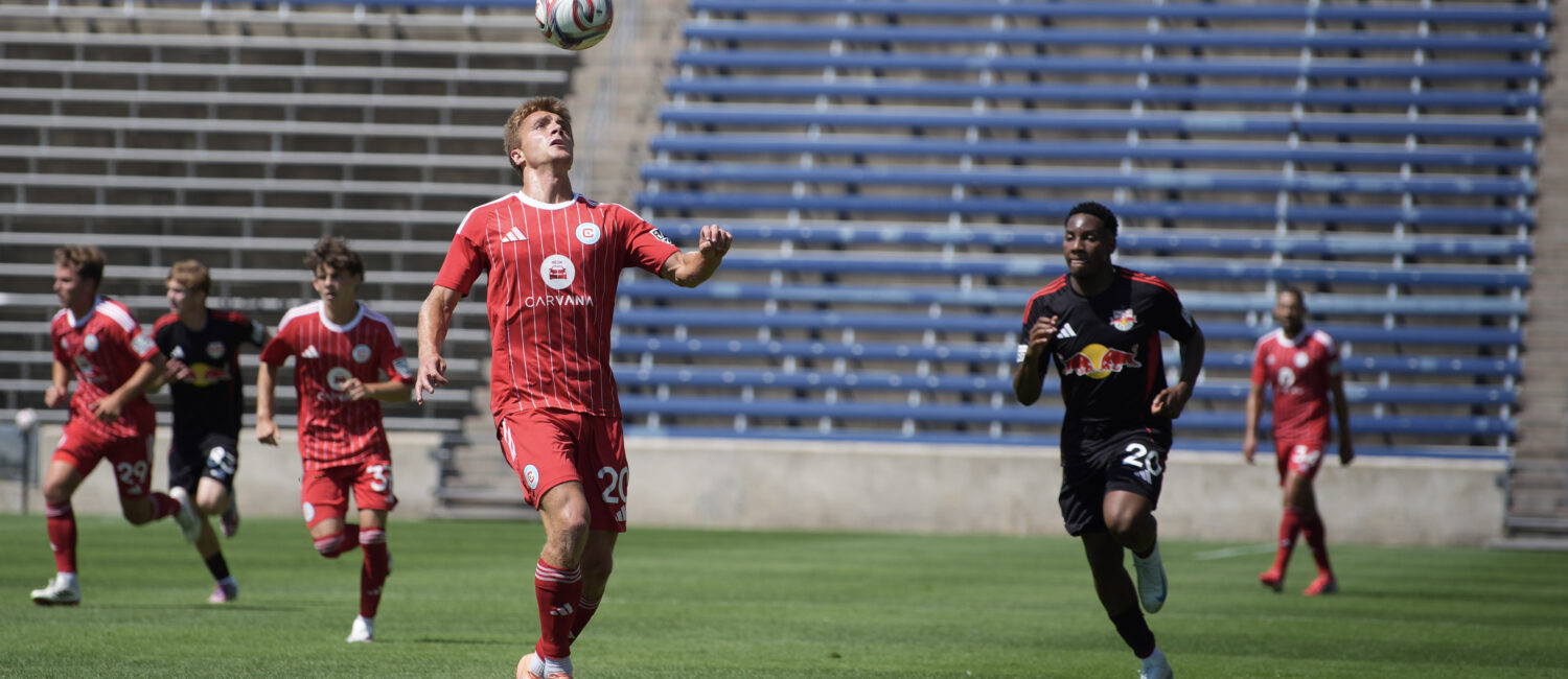 Chicago Fire II player in a red uniform runs toward a ball in the air preparing to take a header, with several opposing players chasing behind him on a field.
