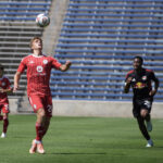 Chicago Fire II player in a red uniform runs toward a ball in the air preparing to take a header, with several opposing players chasing behind him on a field.
