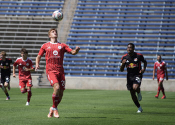 Chicago Fire II player in a red uniform runs toward a ball in the air preparing to take a header, with several opposing players chasing behind him on a field.