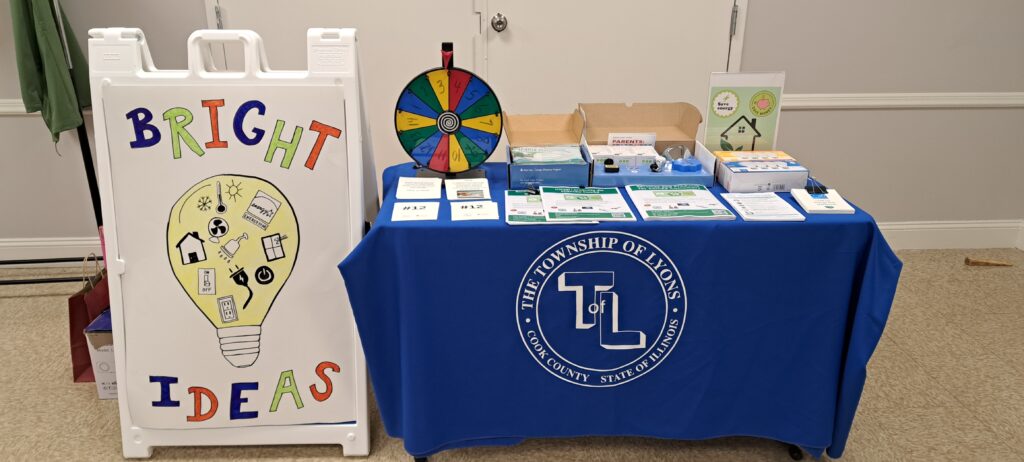 Township of Lyons table with handouts and resources about saving energy. Next to it is a large sign reading "Bright Ideas" with a drawing of a lightbulb and parts of a home that use energy.