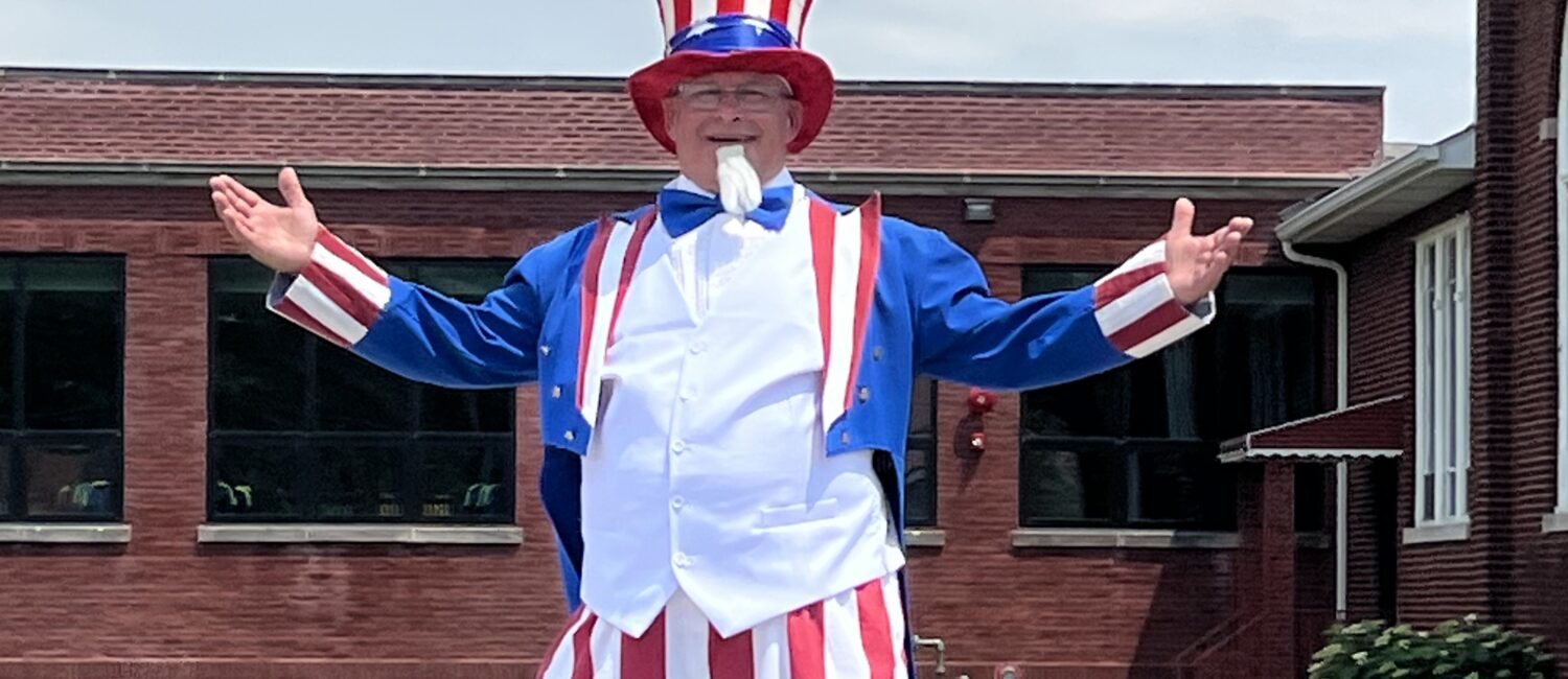 A smiling person dressed as Uncle Sam stands on stilts with their arms outstretched during a Fourth of July parade. He is wearing a tall red, white, and blue striped hat, a blue jacket, a white vest, red-and-white striped pants that cover the stilts. He is in front of a brick building and it is cloudy outside.