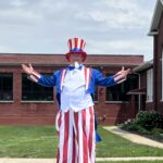 A smiling person dressed as Uncle Sam stands on stilts with their arms outstretched during a Fourth of July parade. He is wearing a tall red, white, and blue striped hat, a blue jacket, a white vest, red-and-white striped pants that cover the stilts. He is in front of a brick building and it is cloudy outside.