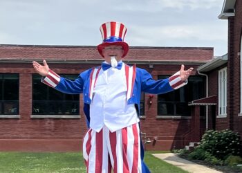A smiling person dressed as Uncle Sam stands on stilts with their arms outstretched during a Fourth of July parade. He is wearing a tall red, white, and blue striped hat, a blue jacket, a white vest, red-and-white striped pants that cover the stilts. He is in front of a brick building and it is cloudy outside.