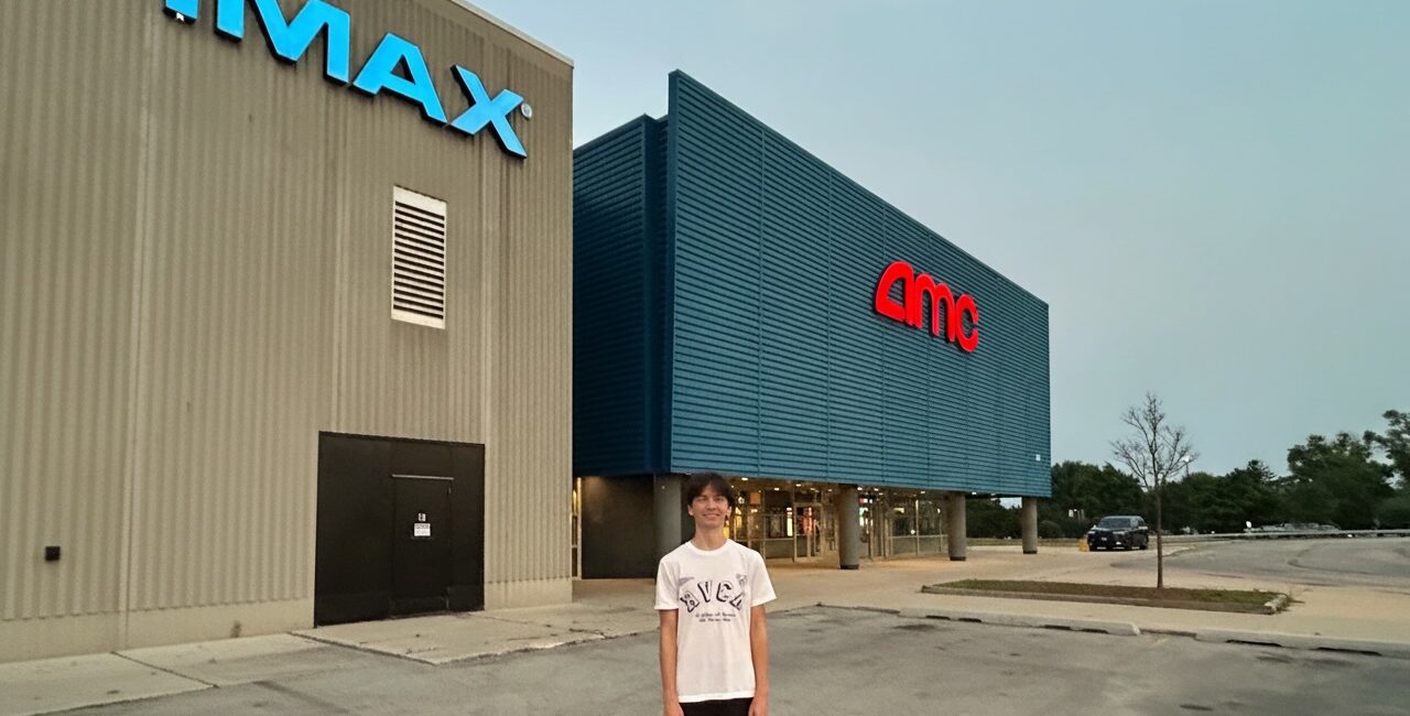 Young man stands in front of an IMAX AMC movie theater on a day with clear skys.