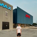 Young man stands in front of an IMAX AMC movie theater on a day with clear skys.