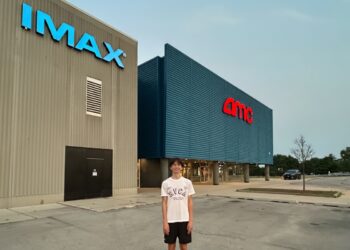 Young man stands in front of an IMAX AMC movie theater on a day with clear skys.