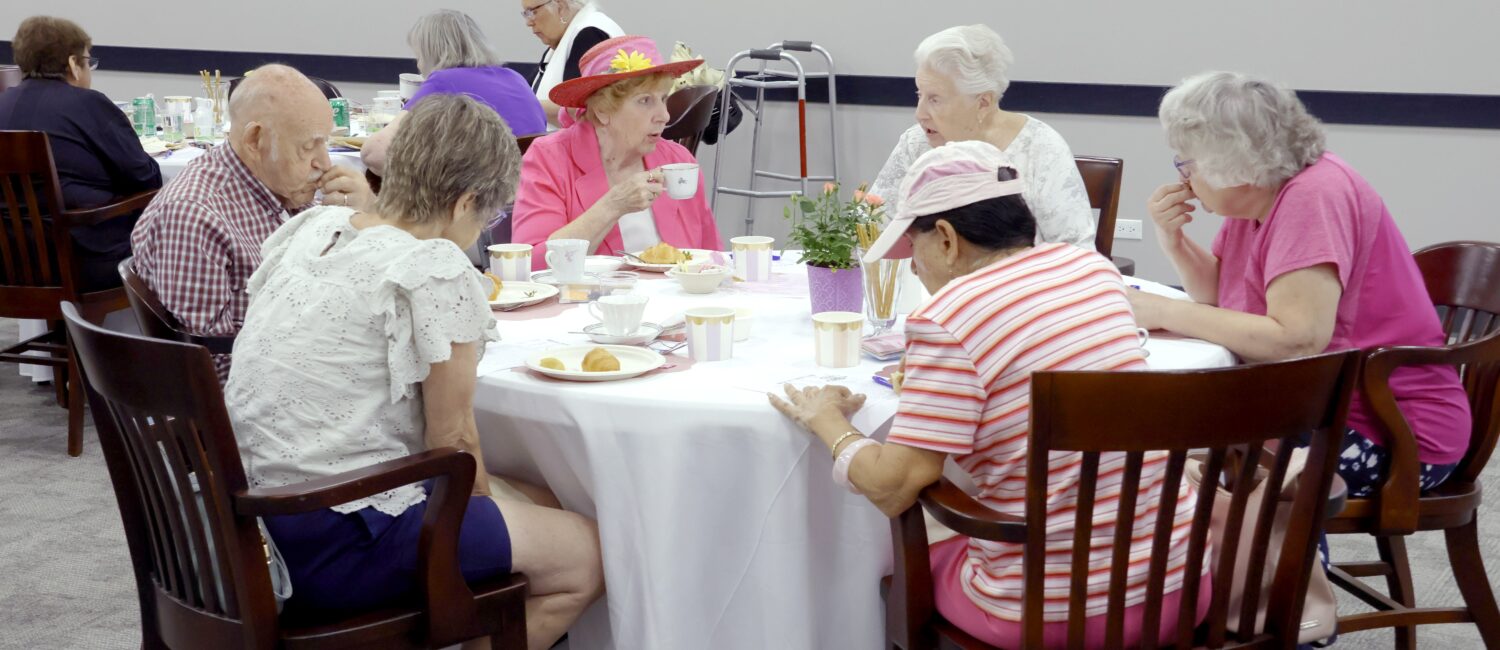A group of older adults gathers around a table for tea and pastries. They sit in a bright community room, with cups, plates and a small floral centerpiece on the table. One person wears a colorful flowered hat, and a walker is visible in the background.