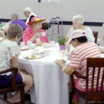 A group of older adults gathers around a table for tea and pastries. They sit in a bright community room, with cups, plates and a small floral centerpiece on the table. One person wears a colorful flowered hat, and a walker is visible in the background.