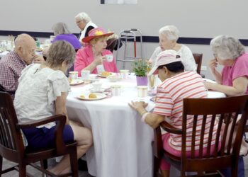 A group of older adults gathers around a table for tea and pastries. They sit in a bright community room, with cups, plates and a small floral centerpiece on the table. One person wears a colorful flowered hat, and a walker is visible in the background.